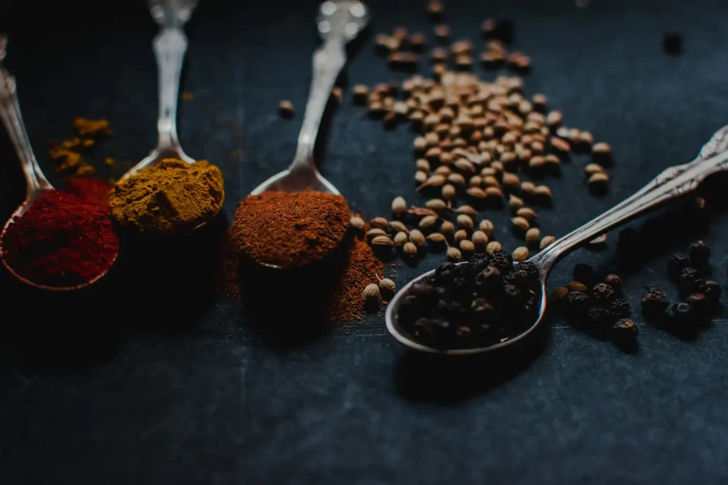 Image of several spoons containing traditional Balinese spices used to make base genep Bali seasoning. The spices include red chili powder, turmeric powder, cinnamon powder, coriander seeds, and black pepper