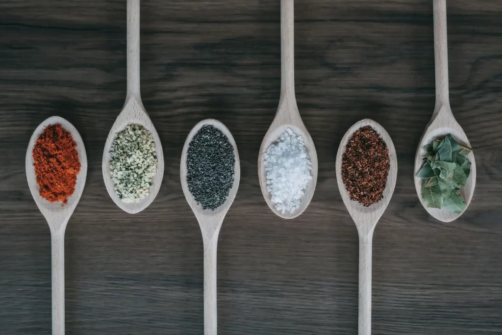 Image of several wooden spoons containing traditional Balinese spices used to make base genep Bali seasoning. The spices include red chili powder, coarse sea salt, and dried chili flakes.